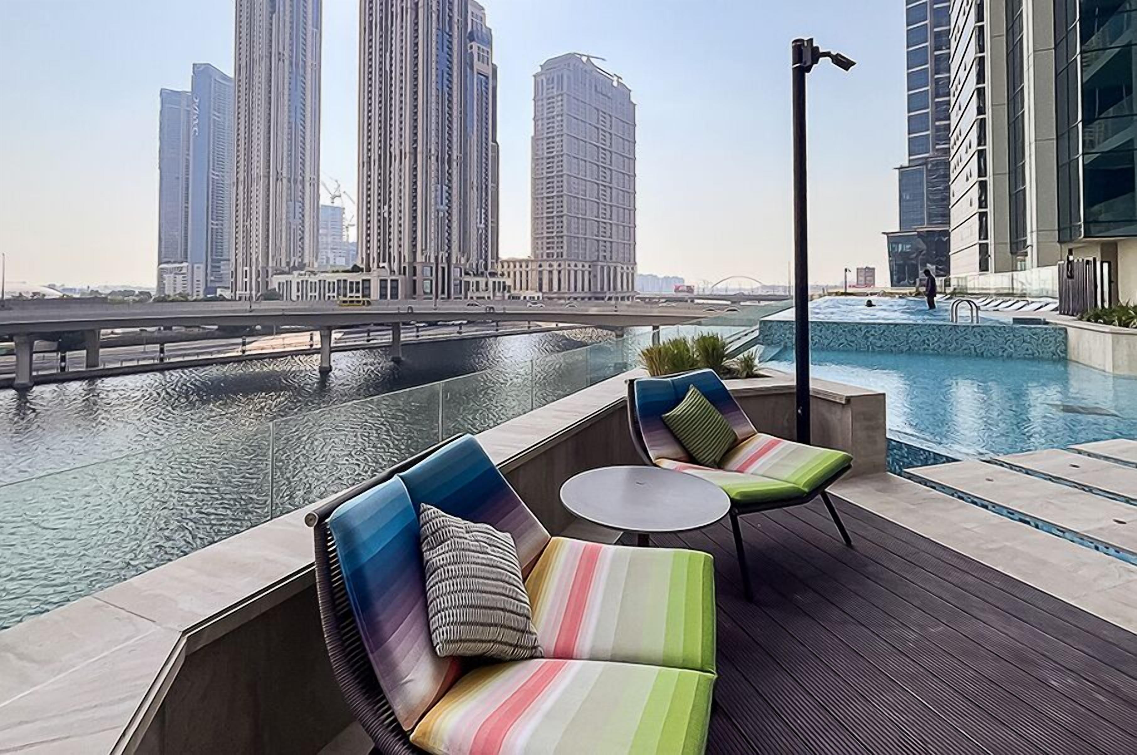 A rooftop pool area with seating and a view of high-rise buildings across a body of water.