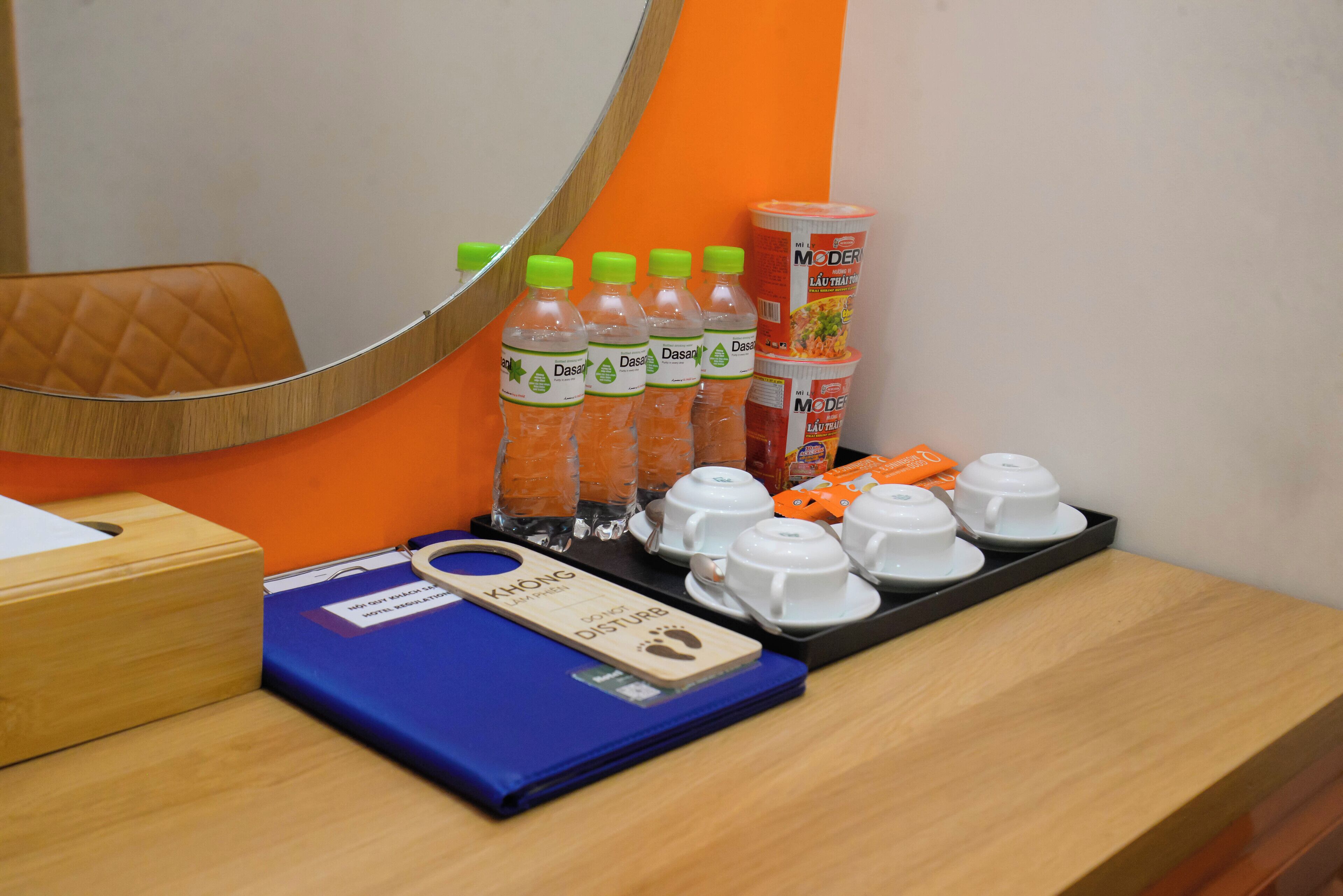 A hotel room with a wooden desk, a blue folder, a wooden footrest, and a tray with cups and bottles.