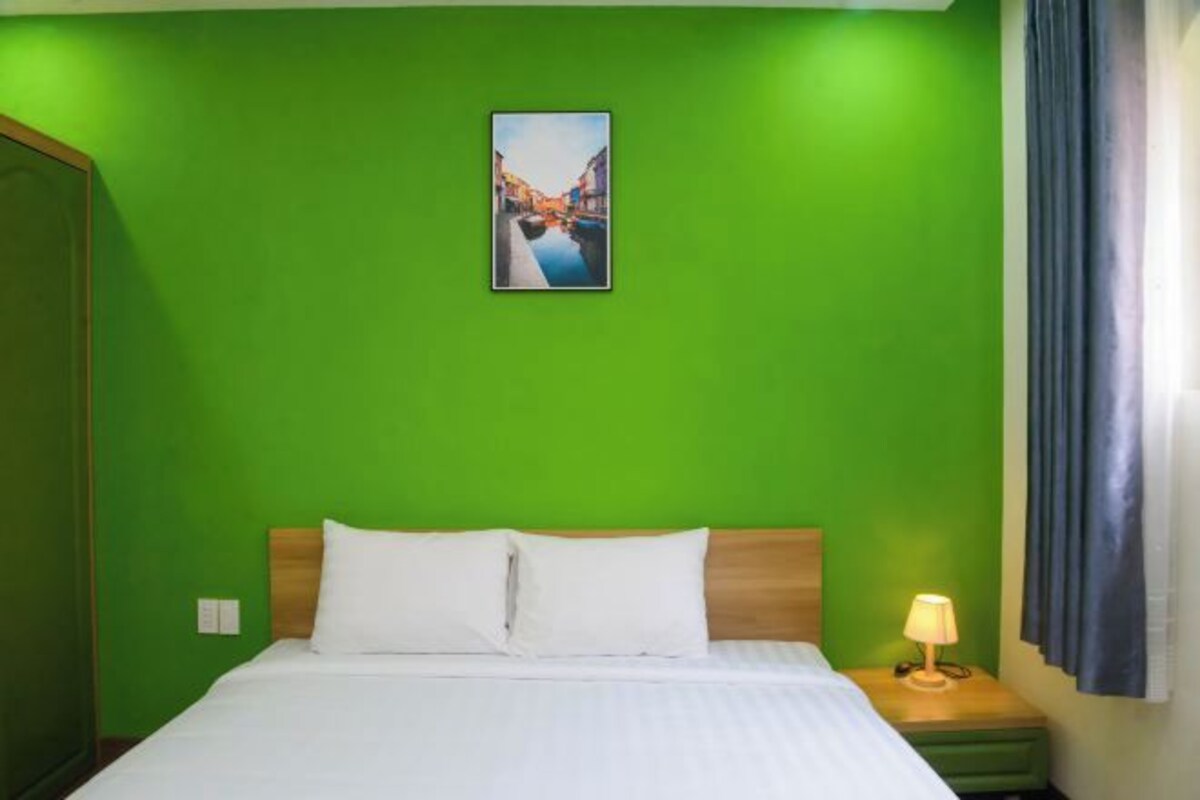 A hotel room with a green wall, a bed with white bedding, a nightstand with a lamp, and a painting of a canal.