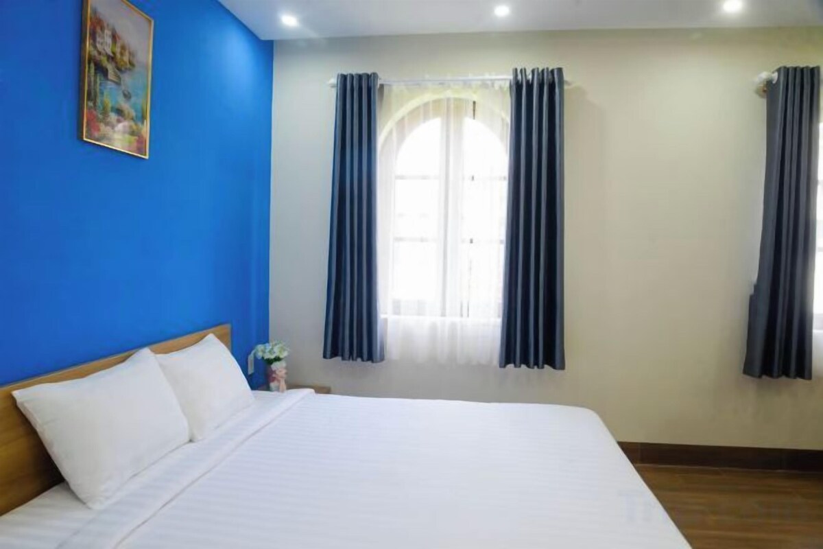 Una habitación de hotel con una cama grande, una mesita de noche, una ventana con cortinas y un cuadro en la pared.