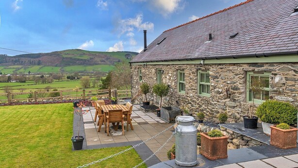 A stone house with a tiled roof, a wooden table and chairs outside, and a view of green fields and hills in the background. - Beautiful holiday cottage in Chittlehampton, No deposit (Llandrillo)