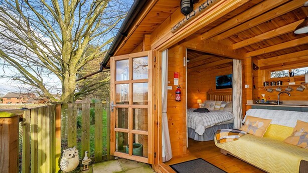 Een gezellig blokhut met houten gevel, een grote openslaande deur naar een slaapkamer met een bed en een bank, en een kleine keukenhoek.