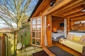 A cozy log cabin with a wooden exterior, a large open door leading to a bedroom with a bed and a sofa, and a small kitchen area. - Pet-friendly chalet in Surlingham, No deposit (Surlingham)