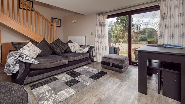 A modern living room with a grey sofa, ottoman, and a desk with a chair. A staircase with wooden railings is visible in the background. - Cottage in Norfolk near River Yare (Surlingham)