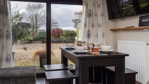 A dining area with a table set for four, a television mounted on the wall, and a view of the garden outside. - Cottage in Norfolk near River Yare (Surlingham)