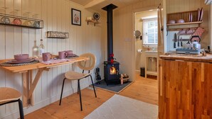 Une cuisine chaleureuse aux murs en bois, avec un poêle à bois central, une table à manger entourée de chaises et des étagères remplies d’ustensiles de cuisine.