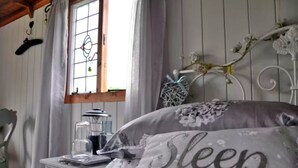 A bedroom with a bed, a nightstand, a window with curtains, and a decorative wall piece. - Shepherd's Hut in Romsey with Fire Pit (Romsey)