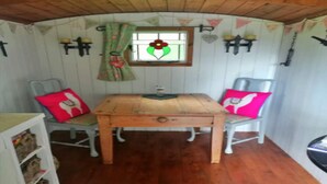 A small room with a wooden table, two chairs, a window with a flower design, and a shelf with baskets. - Shepherd's Hut in Romsey with Fire Pit (Romsey)