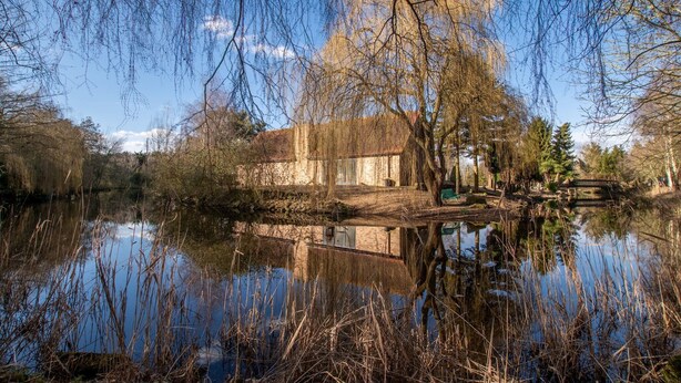 A serene lakeside scene with a rustic building, a wooden bridge, and surrounding trees. - Cottage on Private Island in Cambridgeshire (Ramsey)