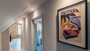 A hallway with a framed poster of a Monaco 1952 race car, a staircase, and a view into a room with a white sink. - Cottage on Private Island in Cambridgeshire (Ramsey)