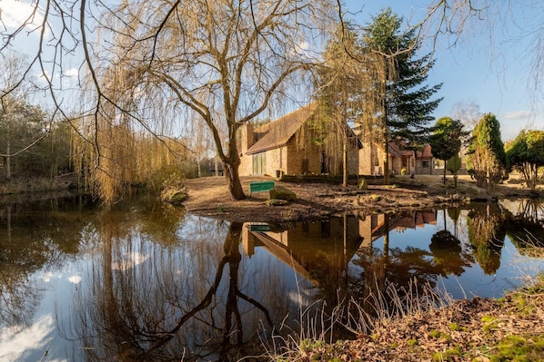 A rustic building with a stone facade, situated by a calm pond reflecting trees and a clear sky.