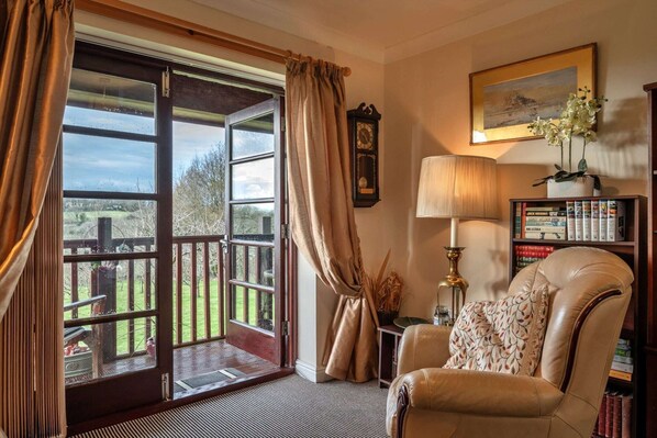 A cozy room with a leather armchair, a bookshelf, a lamp, and a large window with curtains.