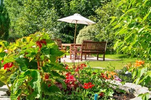 A garden patio with a wooden table and chairs, surrounded by lush greenery and flowering plants.