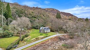 A rural house with a stone exterior, surrounded by a grassy area and trees, with a road leading to it. - Cottage by Loch with Stunning Views (Achahoish)