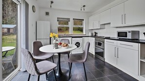 A modern kitchen with white cabinets, a built-in oven, microwave, and washing machine. There is a round table with chairs and a vase of flowers. - Cottage by Loch with Stunning Views (Achahoish)