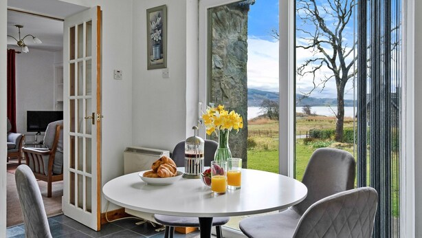 A dining area with a table set for breakfast, including croissants, a vase with flowers, and glasses of orange juice. - Cottage by Loch with Stunning Views (Achahoish)