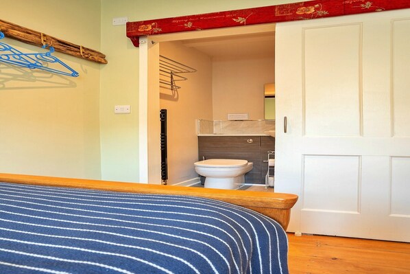 A bedroom with a bed, a wooden headboard, a blue-striped bedspread, a wooden floor, a white door, and a bathroom visible through an open door.
