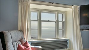 Una habitación con un ventanal grande, un radiador, un sillón gris y una almohada roja.