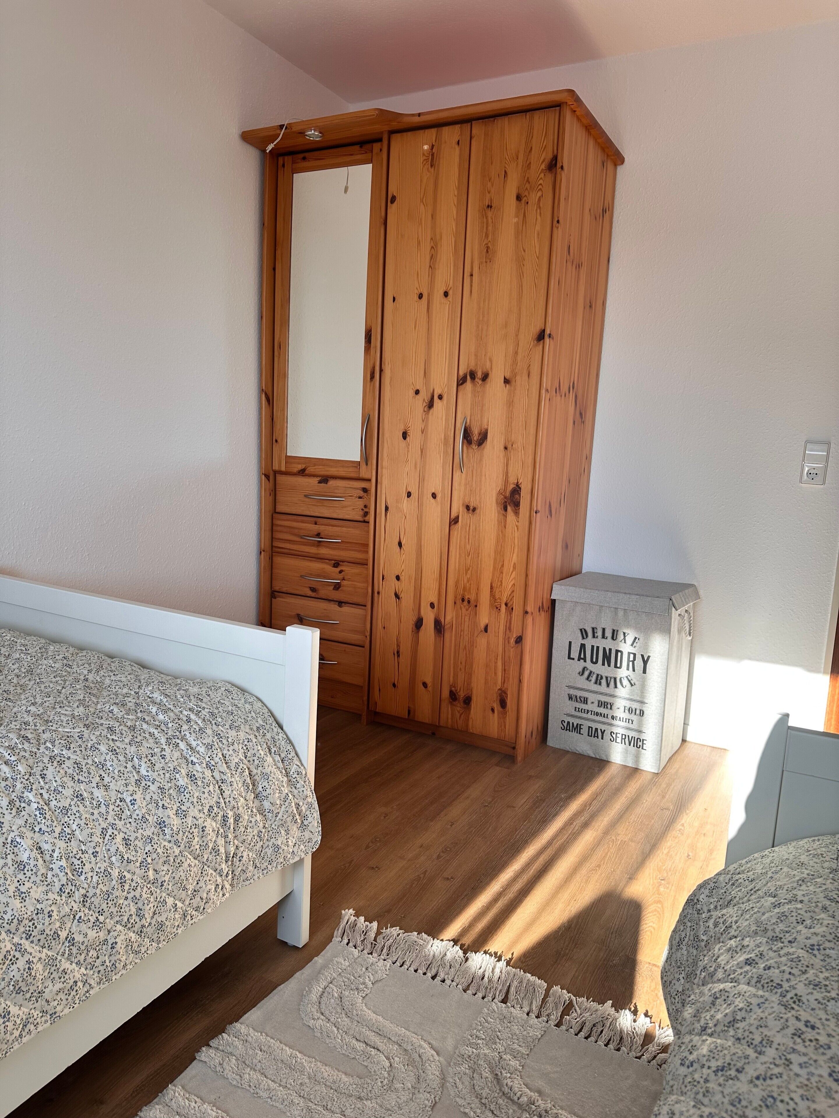 Ein Schlafzimmer mit einem Holzschrank, einem Bett mit floralem Überwurf, einem kleinen Nachttisch und einer Box mit dem Aufdruck 'Deluxe Laundry Service'.