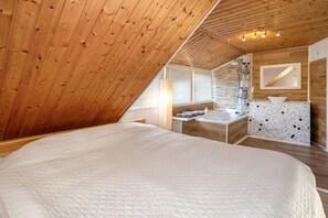 A bedroom with a wooden ceiling, a large bed, a bathroom with a sink and shower, and a mirror.