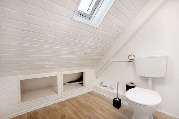 Une salle de bain blanche avec un plafond en pente, une fenêtre, des toilettes et un balai.