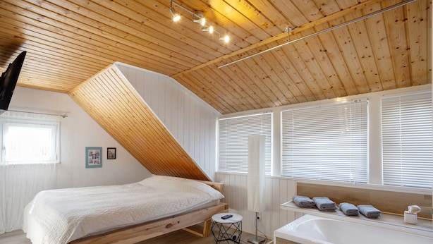 A bedroom with a slanted ceiling, a bed, a bedside table, a lamp, a window with blinds, and a bathroom with a sink and towels. - Holiday Apartment 'Traumhaft' with Shared Garden and Wi-Fi (Wasserburg)
