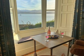 A room with a view of a lake and mountains, a table with a bottle and glasses, and a bench. - Apartment in Beaumaris with Sea Views (Beaumaris)