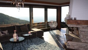 A living room with a sofa, round coffee table, and a chair, large windows with a view, and a fireplace. - Holiday Home 'Talblick' with Mountain View, Private Terrace and Wi-Fi (Sasbachwalden)