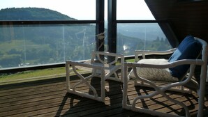 A wooden deck with a white chair and a view of a hilly landscape through large windows. - Holiday Home 'Talblick' with Mountain View, Private Terrace and Wi-Fi (Sasbachwalden)