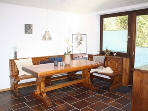 Een eetgedeelte met een houten tafel en bank, een vaas met bloemen, hanglampen en een raam met uitzicht op groen.