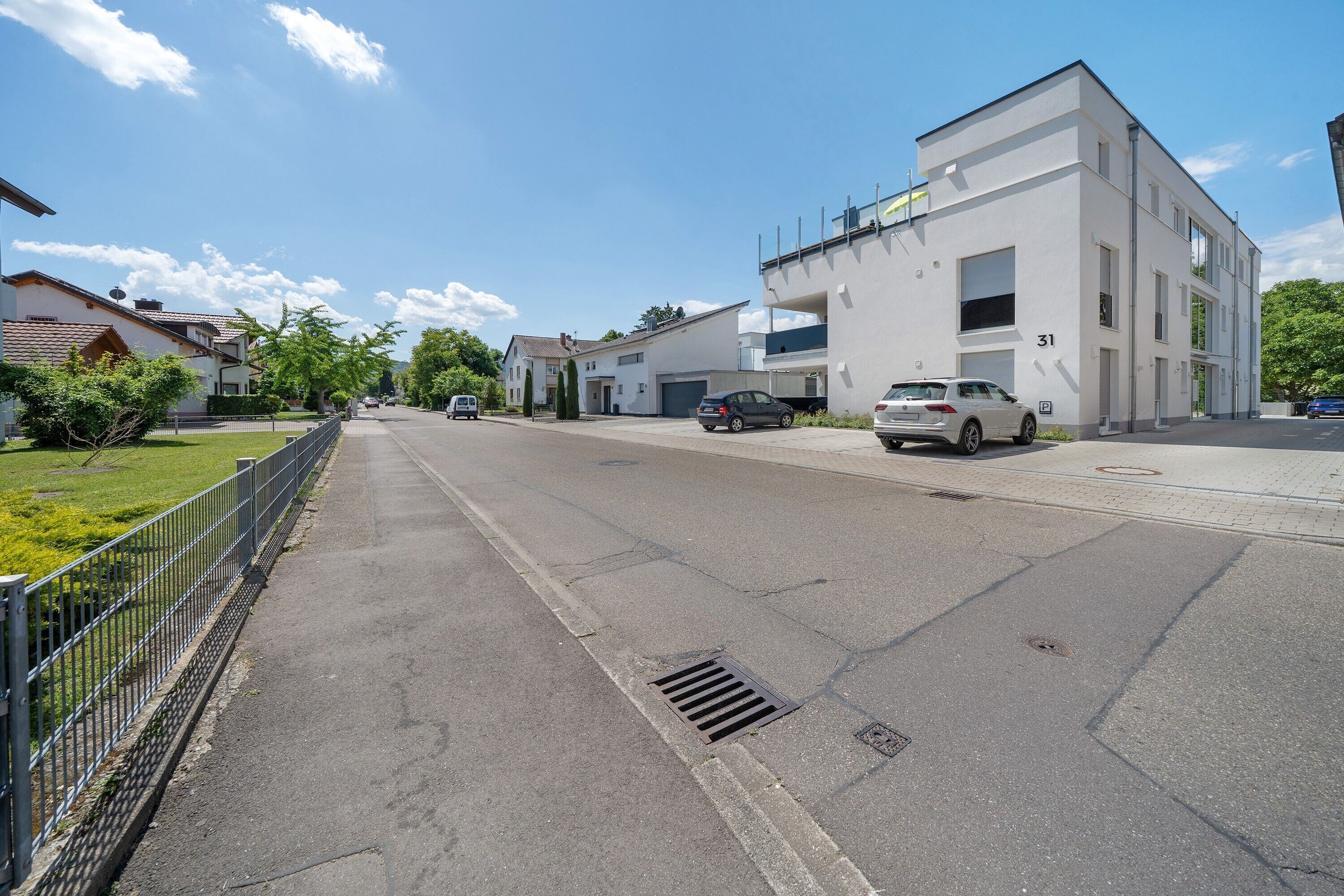 Une rue résidentielle bordée de maisons modernes, avec une allée privée fermée par un portail et des voitures garées.