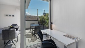 Una habitación moderna con un ventanal que da a un balcón, una mesa blanca, dos sillas y una mesita redonda con un frutero.