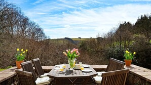An outdoor dining area with a table set for four, surrounded by chairs with striped cushions, a vase with flowers, and potted plants with yellow flowers. - Holiday Home in Mid Wales with Countryside Views (Llanidloes)