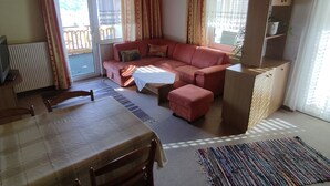 Een gezellige woonkamer met een bank, een voetenbankje en een eettafel met stoelen. Er is een raam met gordijnen, een radiator en een plafondlamp.