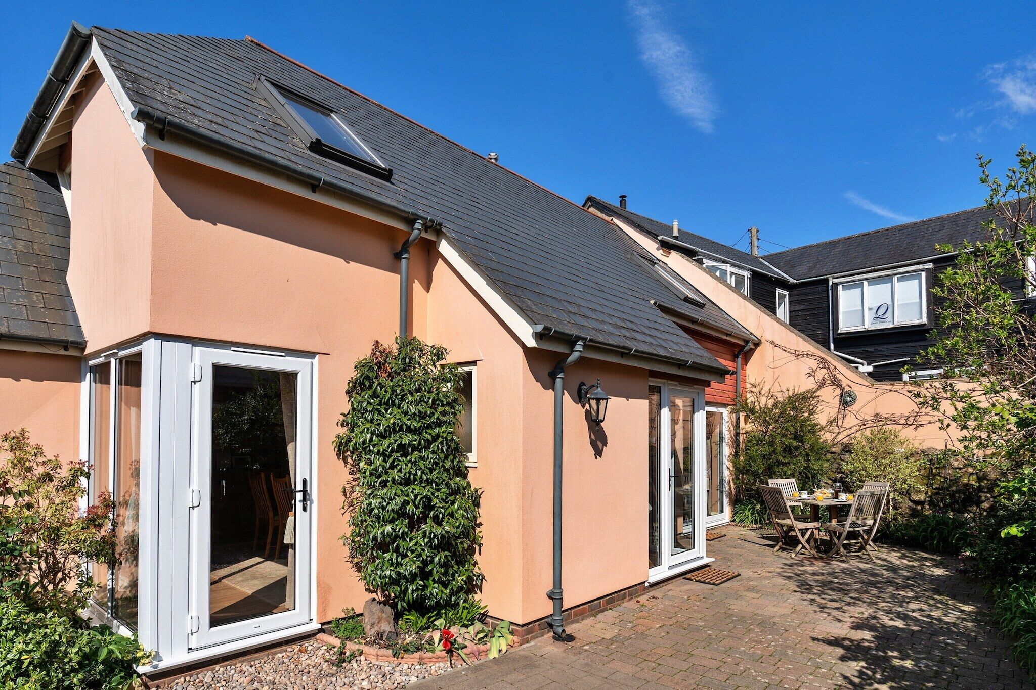 A house with a sloped roof, a sliding glass door, a small patio with a table and chairs, and a garden area with a tall shrub.