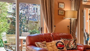 A leather sofa with floral cushions, a wooden table, and chairs outside a sliding glass door. - Cottage in Bovey Tracey near Dartmoor Park (Bovey Tracey)