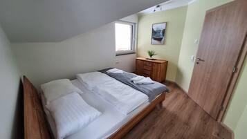 A bedroom with a bed, a wooden wardrobe, a small wooden nightstand, a window, and a painting on the wall.