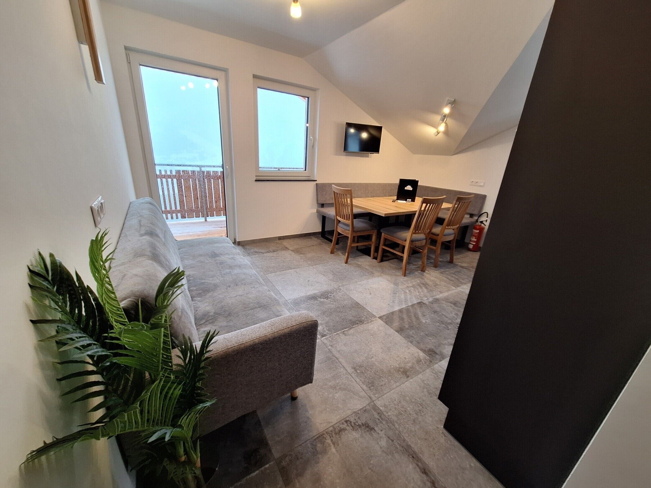 Ein modernes Interieur mit einem grauen Sofa, einem Esstisch mit Stühlen, einem Flachbildfernseher und einem Balkon mit Holzgeländer.