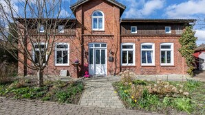 A red brick building with a central entrance, a small garden with plants, and a clear sky. - Apartment 'Fluss Aufwärts' with Private Terrace and Wi-Fi (Schülp)