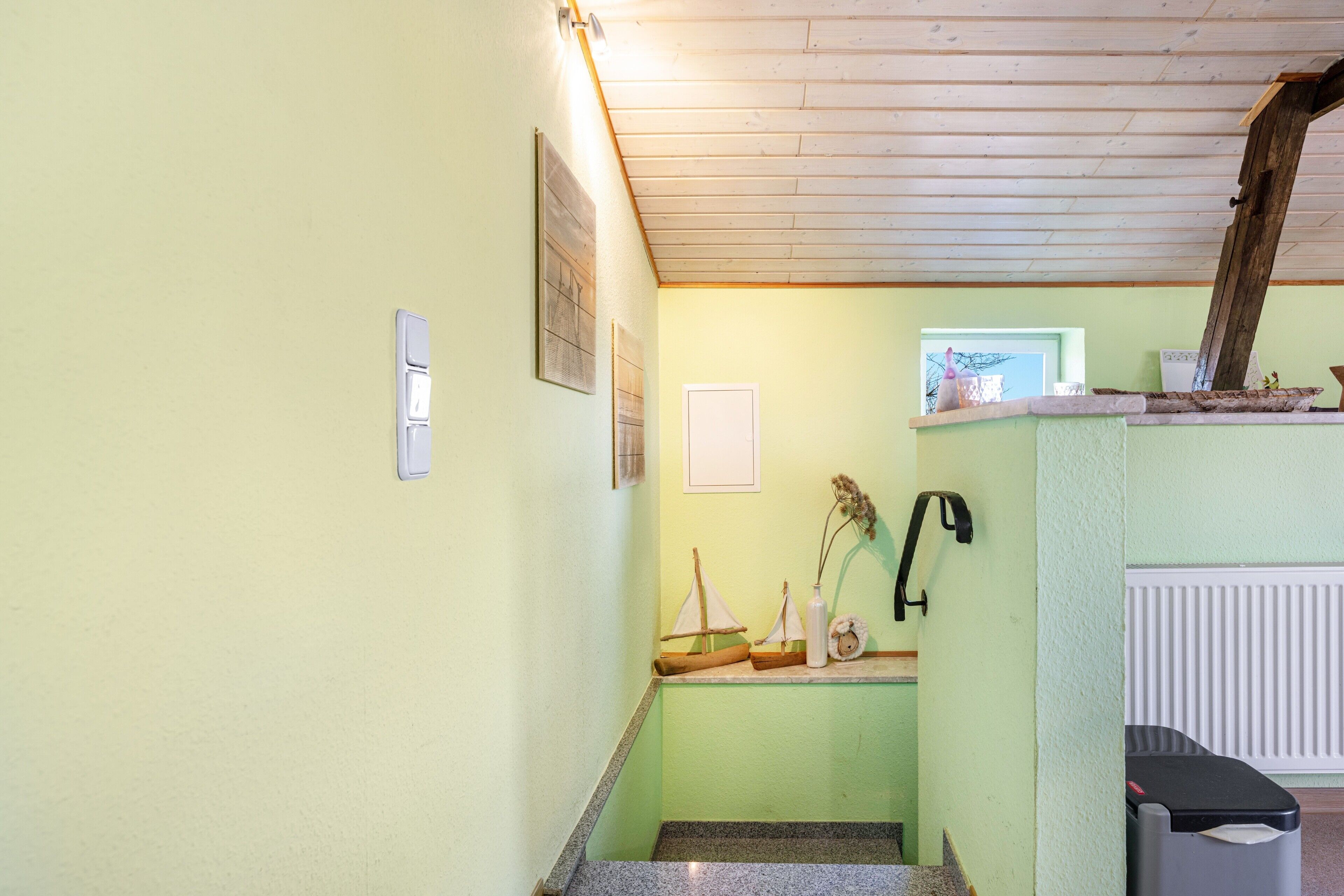 Una habitación con techo de madera, una ventana pequeña, un radiador y un bote de basura.
