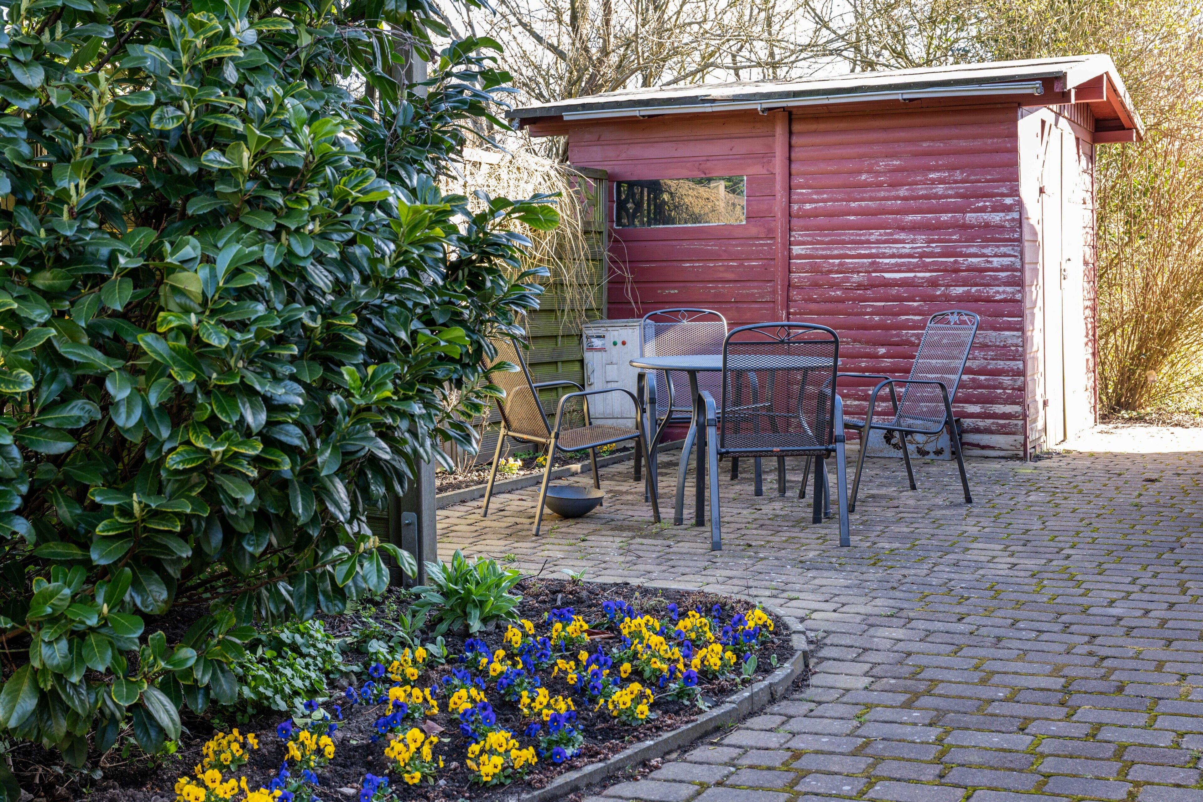 Ein rotes Gartenhaus mit Metallstühlen und einem Tisch, umgeben von einem Garten mit blauen und gelben Blumen.