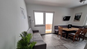 Ett vardagsrum med en soffa, ett matbord med stolar, en krukväxt, en tv och en skjutdörr i glas som leder ut till ett utomhusområde.