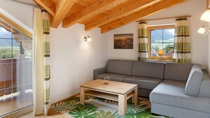 Quarto com teto de madeira, sofá cinza, mesa de centro de madeira e vista pela janela com cortinas.