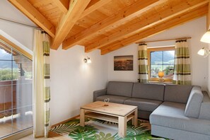 Une pièce avec un plafond en bois, un canapé gris, une table basse en bois et une vue par une fenêtre avec des rideaux.