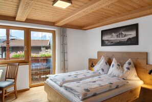 Une chambre avec un plafond en bois, un lit recouvert d’une literie à motifs de feuilles, une chaise, une fenêtre donnant sur des bâtiments et des fleurs, et un tableau encadré accroché au mur.