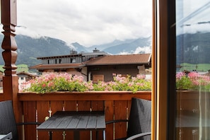 A balcony with a wooden table and chairs, a flower bed with pink flowers, and a view of a mountain range.