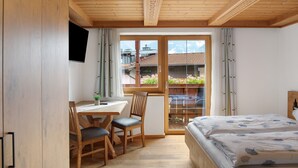 Quarto com teto de madeira, cama com colcha floral, escrivaninha com cadeira e vista para um jardim através de uma porta de vidro.