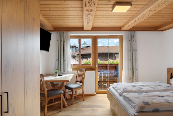 Una habitación con techo de madera, cama con colcha floral, escritorio con silla y vista a un jardín por una puerta de cristal.