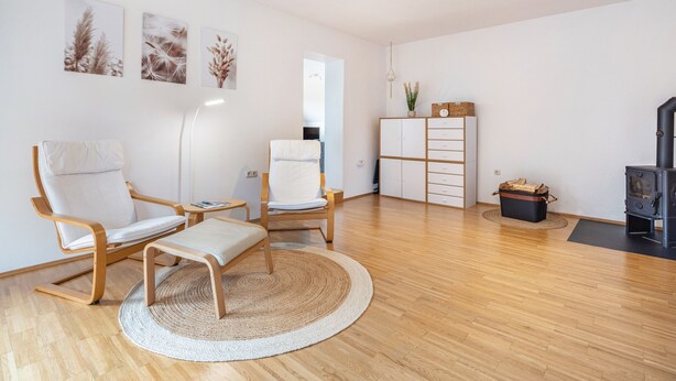 Uma sala de estar moderna com piso de madeira, duas cadeiras brancas, um tapete redondo de palha, um armário e uma lareira.