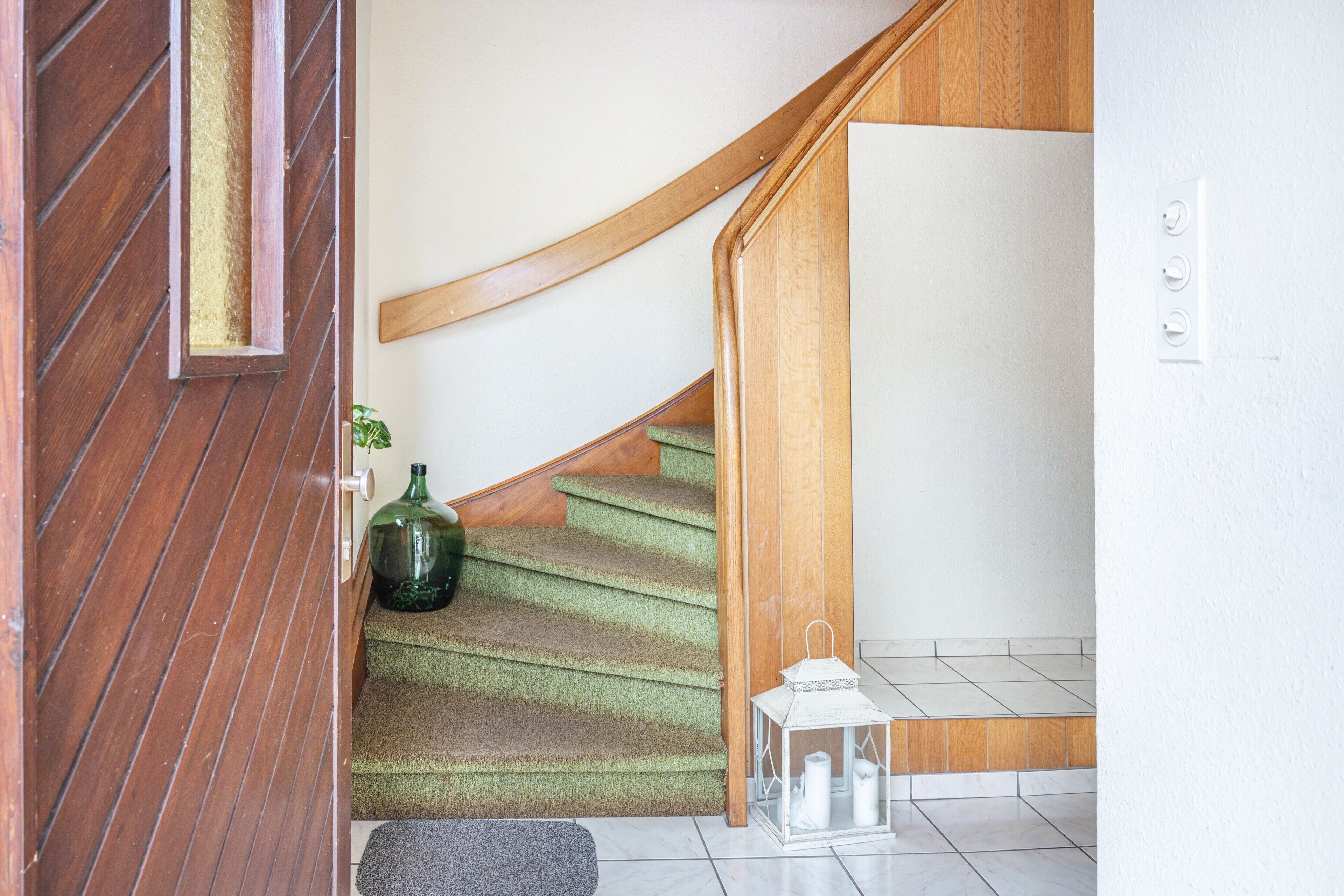 Eine Treppe mit hölzernen Geländern und Teppichstufen, eine grüne Vase und ein weißer Laternenvorbau.